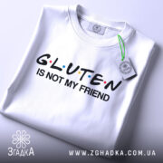 Футболка глютен білого кольору, зображена зверху, на столі, фон світло-сірий, принт чорного кольору з написом 'GLUTEN IS NOT MY FRIEND' та кольоровими крапками, з етикеткою на зеленій стрічці.