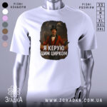 Футболка головний цирк. Біла футболка, зображена з фронтального ракурсу, на манекені. Принт зображає чоловіка в червоному костюмі з капелюхом, який стоїть на фоні циркової арени з акробатами та слоном. Текст на футболці: 'Я керую цим цирком'.