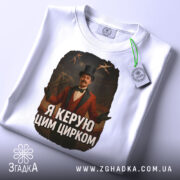 Футболка головний цирк. Біла футболка, складена на поверхні, з видимим етикеткою. Принт зображає чоловіка в червоному костюмі з капелюхом, на фоні циркової арени. Текст на футболці: 'Я керую цим цирком'.