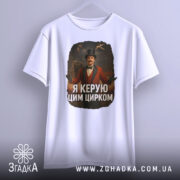 Футболка головний цирк. Біла футболка, зображена з фронтального ракурсу, на манекені. Принт з чоловіком в червоному костюмі з капелюхом, на фоні циркової арени. Текст на футболці: 'Я керую цим цирком'.
