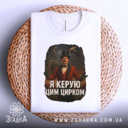 Футболка головний цирк. Біла футболка, складена на круглій поверхні з плетеного матеріалу. Принт з чоловіком в червоному костюмі з капелюхом, на фоні циркової арени. Текст на футболці: 'Я керую цим цирком'.