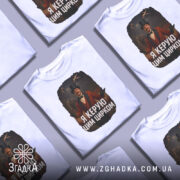 Футболка головний цирк. Біла футболка, зображена з фронтального ракурсу, на манекені. Принт зображає чоловіка в червоному костюмі з капелюхом, який стоїть на фоні циркової арени з акробатами та слоном. Текст на футболці: 'Я керую цим цирком'.