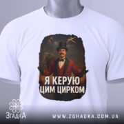 Футболка головний цирк. Біла футболка, складена на поверхні, з видимим етикеткою. Принт зображає чоловіка в червоному костюмі з капелюхом, на фоні циркової арени. Текст на футболці: 'Я керую цим цирком'.