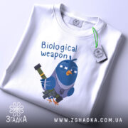 Футболка голуб біологічна зброя, біла, складена на столі, фон світло-сірий, принт з мультяшним птахом у військовому стилі, що тримає зброю, текст 'Biological weapon' над птахом, на етикетці зелений шнурок.