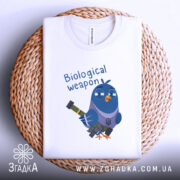 Футболка голуб біологічна зброя, біла, складена на плетеній кошику, фон світло-сірий, принт з мультяшним птахом у військовому стилі, що тримає зброю, текст 'Biological weapon' над птахом.