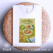 Футболка голубці білого кольору, складена на плетеній кошику, фон світло-сірий, принт з малюнком голубців, овочів та інших страв, оформлений у яскравих кольорах.