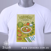 Футболка голубці білого кольору, складена на поверхні, фон світло-сірий, принт з малюнком голубців, овочів та інших страв, прикрашений етикеткою з зеленим шнурком.