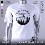 Футболка гори кличуть, біла, зображена з фронтального ракурсу, на фоні світло-сірого градієнту, принт з чорними горами та лісом, текст українською мовою.