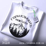Футболка гори кличуть, біла, зображена зверху, на фоні світло-блакитного кольору, принт з чорними горами та лісом, з зеленим етикеткою.