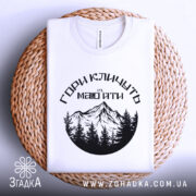 Футболка гори кличуть, біла, зображена зверху на плетеному кошику, фон білий, принт з чорними горами та лісом, текст українською мовою.
