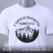 Футболка гори кличуть, біла, зображена зверху, на фоні світло-блакитного кольору, принт з чорними горами та лісом, з зеленим етикеткою.