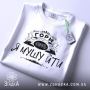 Футболка гори кличуть, білого кольору, складена на поверхні, фон світло-сірий, принт чорного кольору з написом 'ГОРИ КЛИЧУТЬ Я МУШУ ЙТИ' та зображенням гір, з зеленим ярликом.