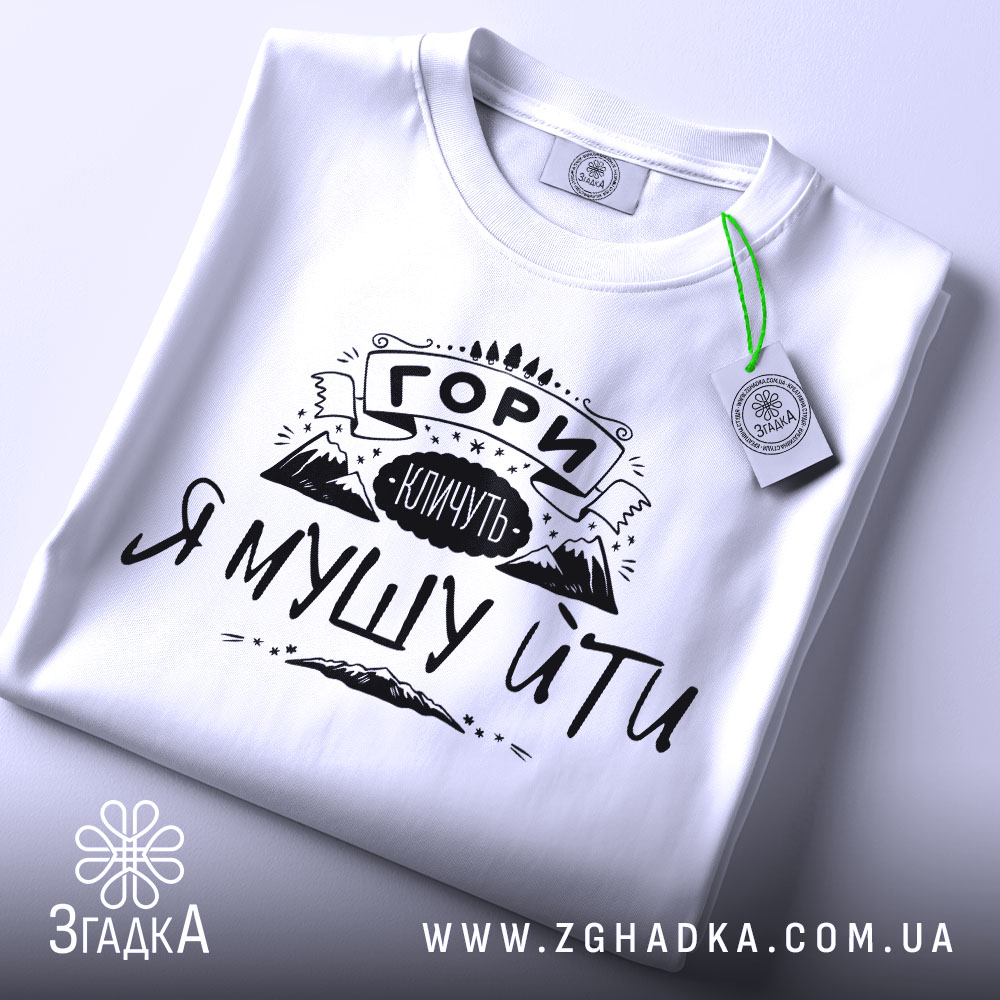 Футболка гори кличуть, білого кольору, складена на поверхні, фон світло-сірий, принт чорного кольору з написом 'ГОРИ КЛИЧУТЬ Я МУШУ ЙТИ' та зображенням гір, з зеленим ярликом.