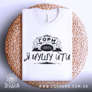 Футболка гори кличуть, білого кольору, складена на плетеній основі, фон світло-сірий, принт чорного кольору з написом 'ГОРИ КЛИЧУТЬ Я МУШУ ЙТИ' та зображенням гір.