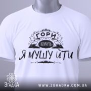 Футболка гори кличуть, білого кольору, складена на поверхні, фон світло-сірий, принт чорного кольору з написом 'ГОРИ КЛИЧУТЬ Я МУШУ ЙТИ' та зображенням гір, з зеленим ярликом.