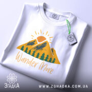 Футболка гори подорожі: біла футболка, складена на столі, з принтом гір, сонця та дерев, текст 'Wander More' у жовтому кольорі, з етикеткою, фон світло-сірий.