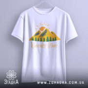 Футболка гори подорожі: біла футболка, підвішена на вішалці, з принтом гір, сонця та дерев, текст 'Wander More' у жовтому кольорі, фон світло-сірий.