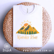 Футболка гори подорожі: біла футболка, складена на плетеній основі, з принтом гір, сонця та дерев, текст 'Wander More' у жовтому кольорі, фон світло-сірий.