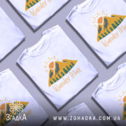 Футболка гори подорожі: біла футболка з принтом, що зображує гори, сонце та дерева, з текстом 'Wander More' у жовтому кольорі, зображена на манекені з металевим блиском, фон світло-сірий.