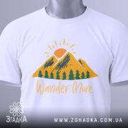 Футболка гори подорожі: біла футболка, складена на столі, з принтом гір, сонця та дерев, текст 'Wander More' у жовтому кольорі, з етикеткою, фон світло-сірий.