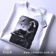 Футболка Горила Діджей білого кольору, складена на плоскій поверхні, фон світло-сірий, принт з горилою в навушниках, що працює на пульті, з пташками на фоні, з етикеткою зеленого кольору.