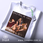 Футболка гострий фемсил білого кольору, складена на поверхні, фон світло-сірий, принт з написом 'GIRLS WILL BE GIRLS' на картині, з зеленим ярликом, що звисає.