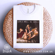 Футболка гострий фемсил білого кольору, складена на плетеній кошику, фон світло-сірий, принт з написом 'GIRLS WILL BE GIRLS' на картині, що зображує жінок у класичному стилі.