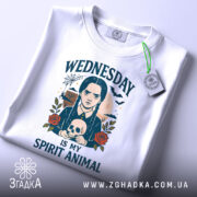 Футболка Готичний Принт, біла, зображена зверху, фон світло-сірий, принт з дівчиною з косами, яка тримає череп, текст 'WEDNESDAY IS MY SPIRIT ANIMAL', на футболці є етикетка з зеленим шнурком.