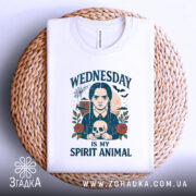 Футболка Готичний Принт, біла, зображена на плетеній кошику, фон світло-сірий, принт з дівчиною з косами, яка тримає череп, текст 'WEDNESDAY IS MY SPIRIT ANIMAL', футболка акуратно складена.