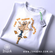 Футболка гра за правилами, біла, складена на поверхні, фон світлий, принт з рукою, що тримає шахову фігуру, з чорними нігтями та жовтим написом, з етикеткою на зеленому шнурку.