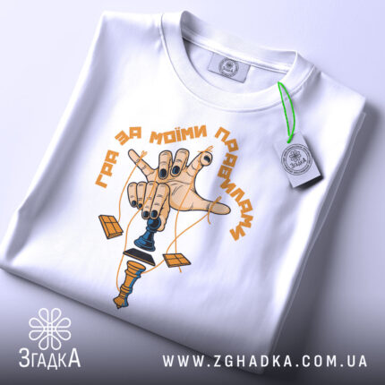 Футболка гра за правилами, біла, складена на поверхні, фон світлий, принт з рукою, що тримає шахову фігуру, з чорними нігтями та жовтим написом, з етикеткою на зеленому шнурку.