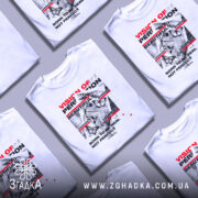 Футболка графічний хаос, біла, зображена на манекені з блискучою поверхнею, фон світло-сірий, принт містить текст у червоному та чорному кольорах, зображення метеликів і графічні елементи.