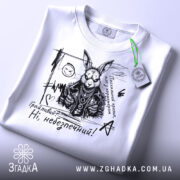 Футболка грайливий небезпечний, білого кольору, зображена зверху, на складеній футболці, фон світло-сірий, принт з чорним малюнком зайця в шкіряному жакеті з написом 'Грайливий? Ні, небезпечний!', з зеленим ярликом.