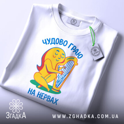 Футболка граю на нервах, біла, складена на столі, фон світло-сірий, принт з жовтою фігурою, що грає на арфі, текст українською мовою 'Чудово граю на нервах', з етикеткою зеленого кольору.