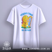 Футболка граю на нервах, біла, підвішена на вішалці, фон світло-сірий, принт з жовтою фігурою, що грає на арфі, текст українською мовою 'Чудово граю на нервах'.