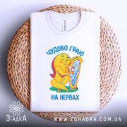 Футболка граю на нервах, біла, складена на плетеній кошику, фон світло-сірий, принт з жовтою фігурою, що грає на арфі, текст українською мовою 'Чудово граю на нервах'.