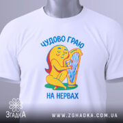 Футболка граю на нервах, біла, складена на столі, фон світло-сірий, принт з жовтою фігурою, що грає на арфі, текст українською мовою 'Чудово граю на нервах', з етикеткою зеленого кольору.