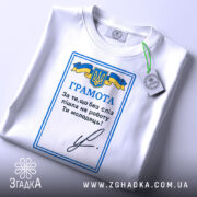 Футболка грамота, біла, складена на столі, фон світлий, принт з синьо-жовтим гербом України та текстом "ГРАМОТА", на комірці є етикетка.