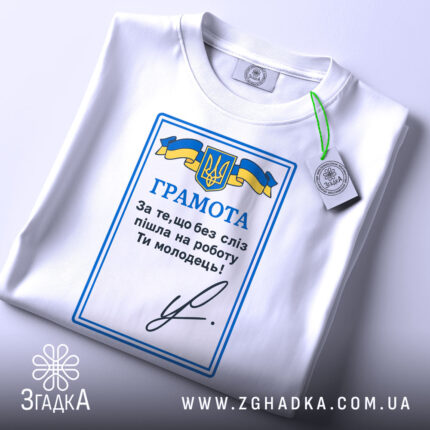 Футболка грамота, біла, складена на столі, фон світлий, принт з синьо-жовтим гербом України та текстом "ГРАМОТА", на комірці є етикетка.