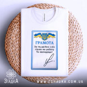 Футболка грамота, біла, складена на плетеному кошику, фон світлий, принт з синьо-жовтим гербом України та текстом "ГРАМОТА".