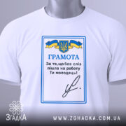 Футболка грамота, біла, складена на столі, фон світлий, принт з синьо-жовтим гербом України та текстом "ГРАМОТА", на комірці є етикетка.