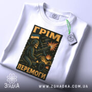 Футболка грім перемоги. Біла футболка, складена на столі, з видимим етикеткою. Принт з військовим персонажем, що стріляє з гармати, на фоні вибухів і українського прапора.