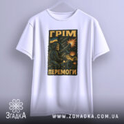 Футболка грім перемоги. Біла футболка з коротким рукавом, підвішена на вішалці, фон світло-сірий. Принт з військовим персонажем, що стріляє з гармати, на фоні вибухів і українського прапора.