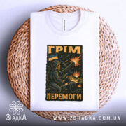 Футболка грім перемоги. Біла футболка, складена на плетеному кошику, фон світло-сірий. Принт з військовим персонажем, що стріляє з гармати, на фоні вибухів і українського прапора.