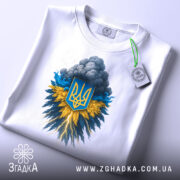 Футболка Грім Тризуб білого кольору, зображена з верхнього ракурсу, на столі з білим фоном, принт з тризубом на синьо-жовтому фоні, з етикеткою на комірці.