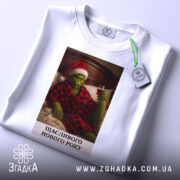 Футболка грінч настрій, складена біла футболка з принтом, на якому грінч у червоному капелюсі та халаті, тримає келих вина, фон - світлий, з легким відблиском, текст під принтом: 'ЩАСЛИВОГО НОВОГО РОКУ'.