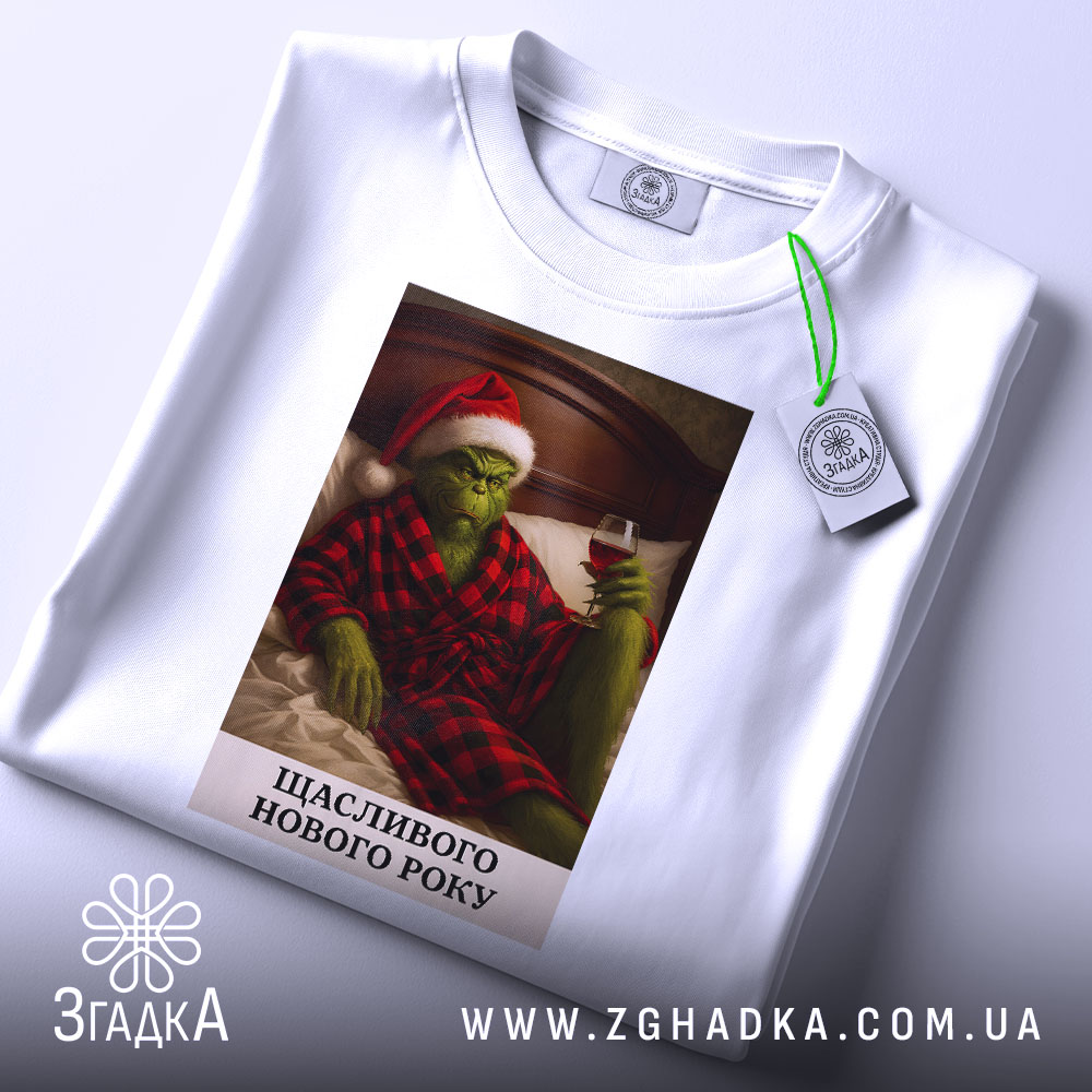 Футболка грінч настрій, складена біла футболка з принтом, на якому грінч у червоному капелюсі та халаті, тримає келих вина, фон - світлий, з легким відблиском, текст під принтом: 'ЩАСЛИВОГО НОВОГО РОКУ'.