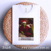 Футболка грінч настрій, складена біла футболка з принтом, на якому грінч у червоному капелюсі та халаті, тримає келих вина, фон - плетений кошик, текст під принтом: 'ЩАСЛИВОГО НОВОГО РОКУ'.