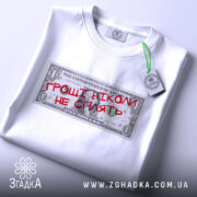 Футболка гроші не сплять, біла, складена на столі, фон світлий, принт у вигляді доларової банкноти з червоним написом "ГРОШІ НІКОМІ НЕ СПІЛЯТЬ", на комірці є етикетка.