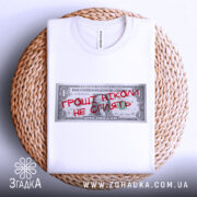 Футболка гроші не сплять, біла, складена на плетеному кошику, фон нейтральний, принт у вигляді доларової банкноти з червоним написом "ГРОШІ НІКОМІ НЕ СПІЛЯТЬ".